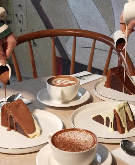 Kek dilimlerinin üzerine çikolata sosu dökülürken çekilmiş bir kare, yanında latte ve üzerine kakao serpilmiş bir kahve