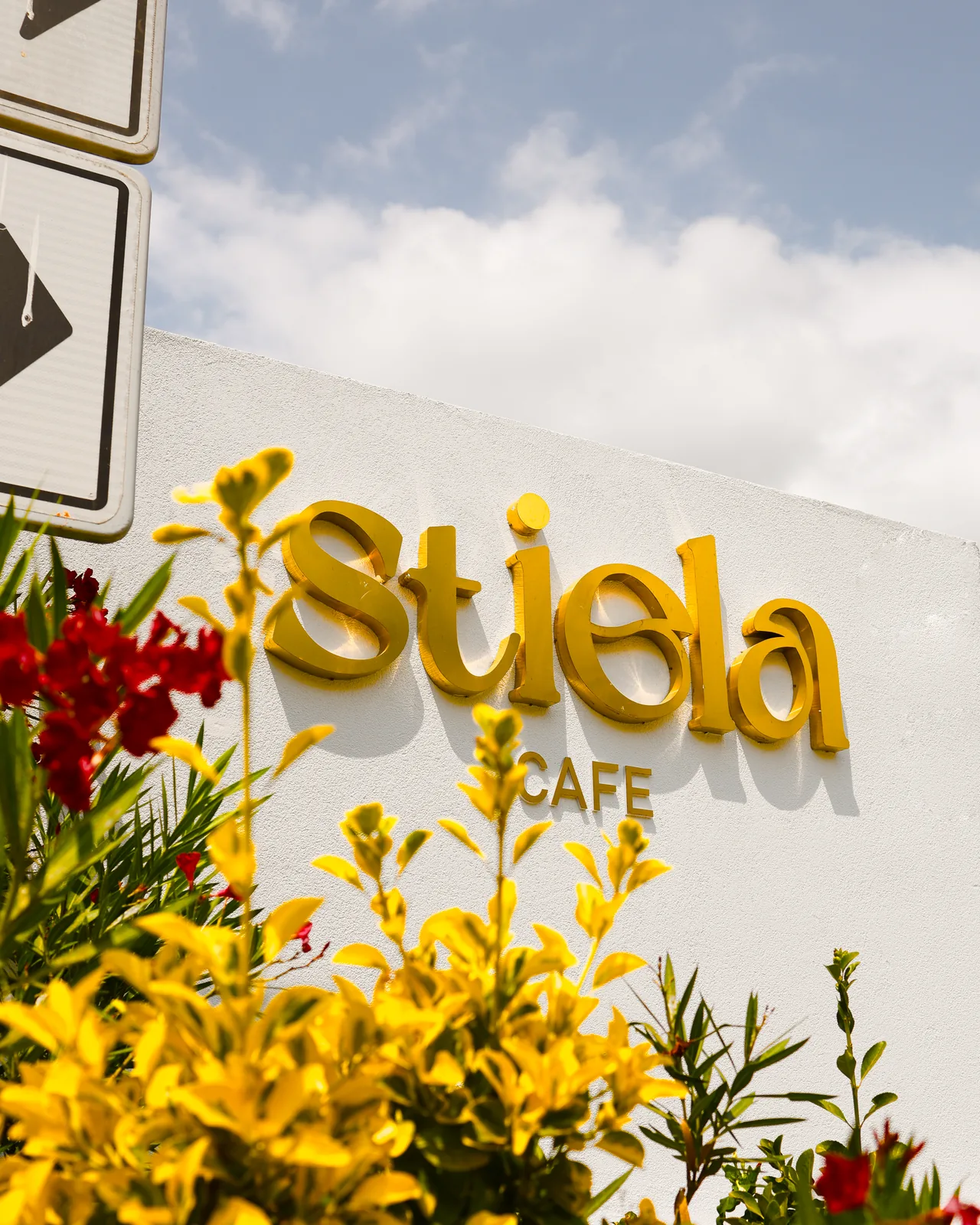 Stiela Cafe’nin beyaz duvardaki sarı harfli tabelası, önünde sarı ve kırmızı çiçekler