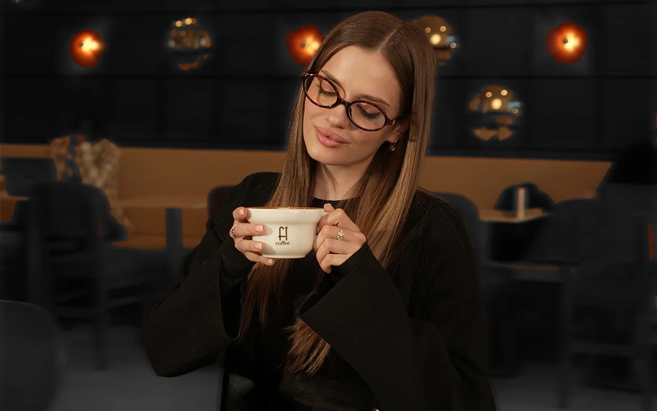 A woman in glasses sipping from a Fi Coffee cup in a softly lit cafe